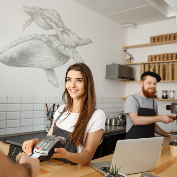 Coffee Business Concept - Beautiful female barista giving payment service for customer with credit card and smiling while working at the bar counter in modern coffee shop.